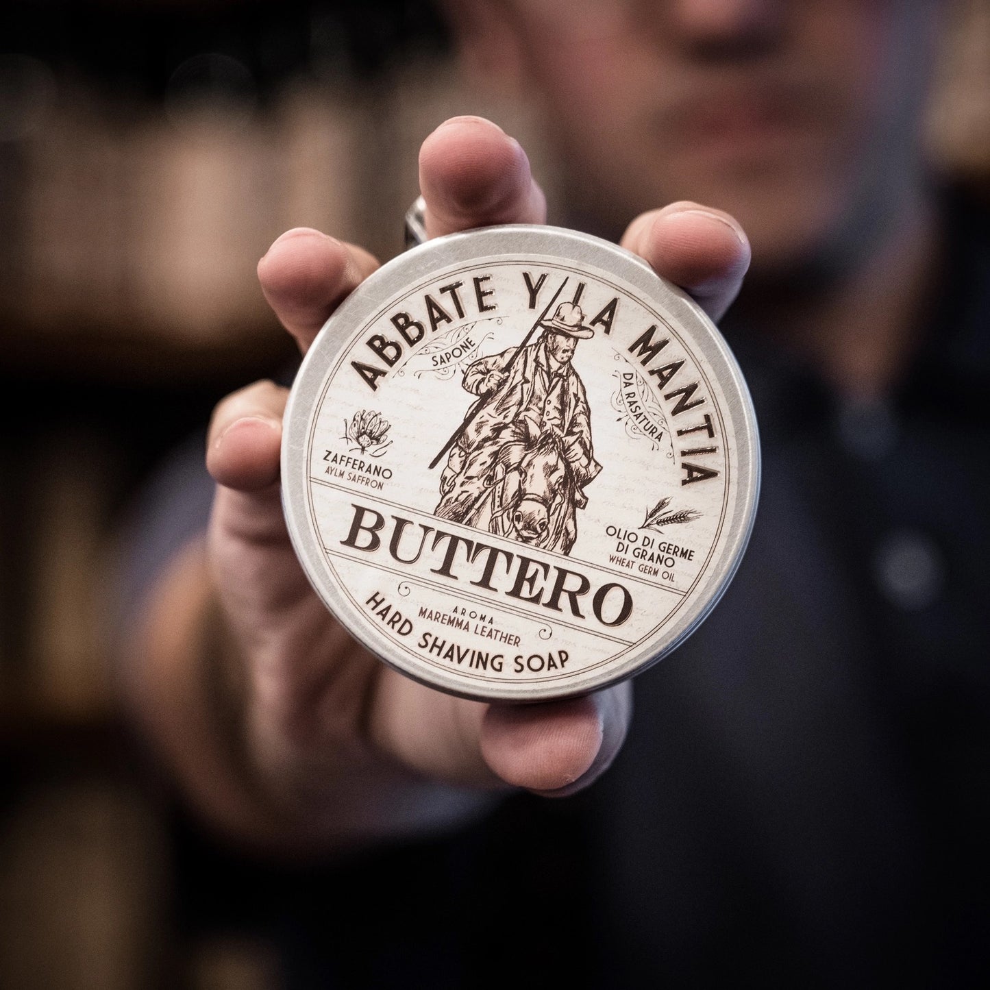 Hand holding a container of Abbate y La Manzia Buttero hard shaving soap with a blurred background.