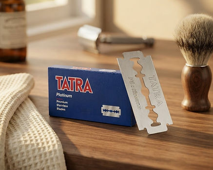 A box of Tatra Platinum disposable razors sits on a wooden table next to a silver razor blade, with a shaving brush and a towel nearby in a warm, sunlit setting.