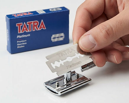 A hand holds a silver Tatra Platinum disposable razor blade over its chrome mounting base, with the blue Tatra product box visible in the background on a white surface.